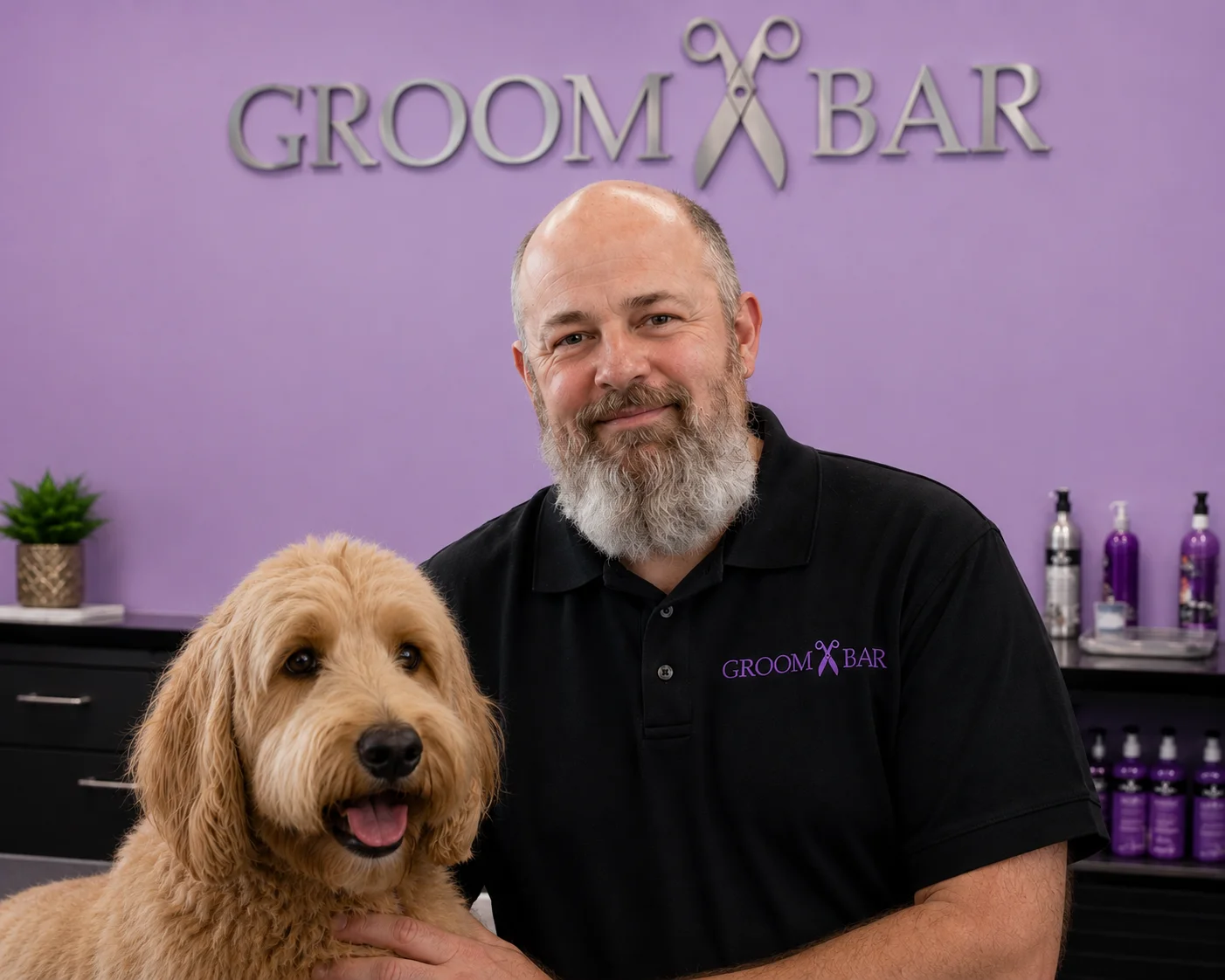 Groombar team member in branded polo posing with a golden doodle in front of the silver scissors logo wall