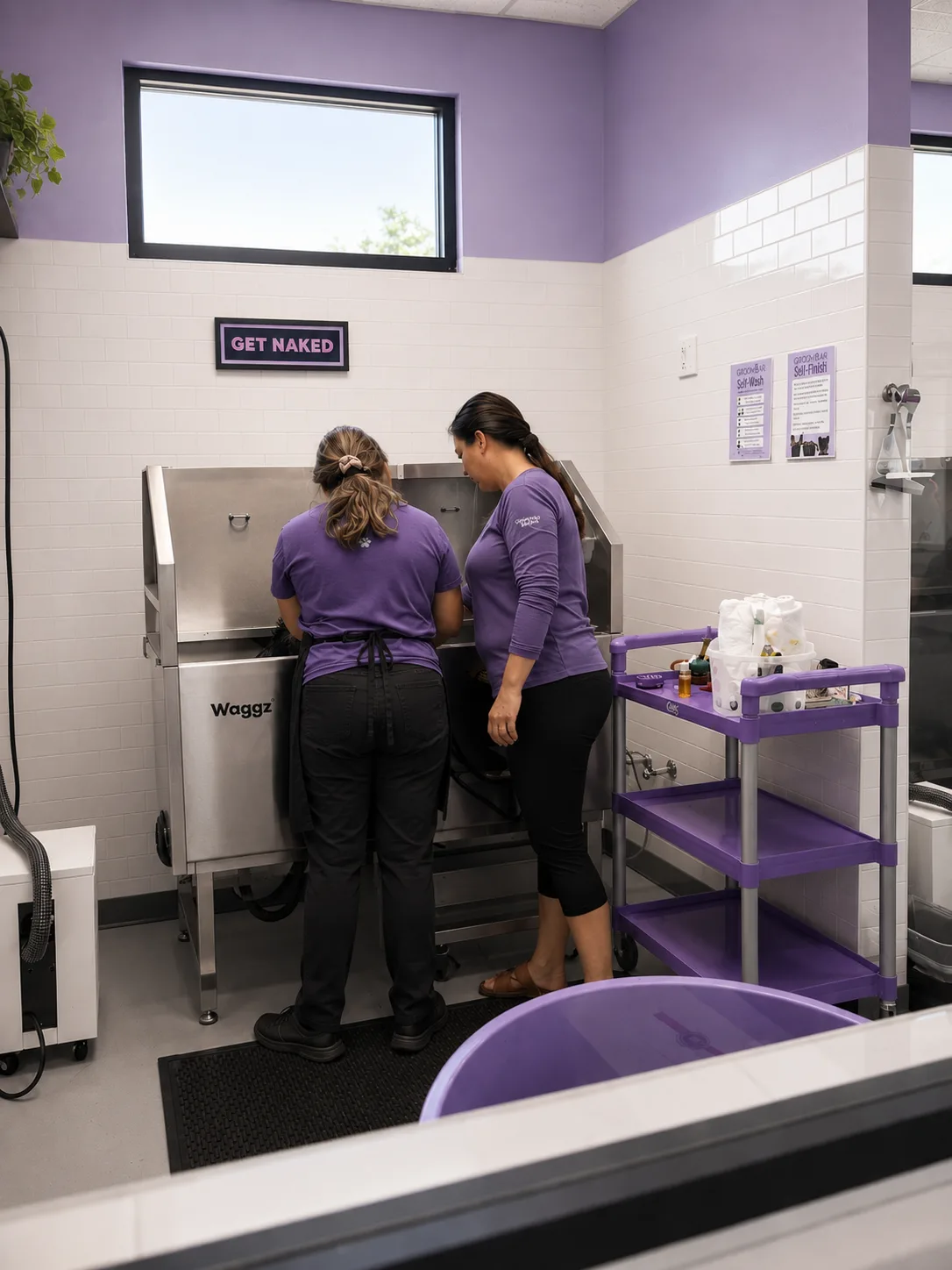 Two Groombar groomers in purple uniforms working together at a stainless steel Waggz wash tub